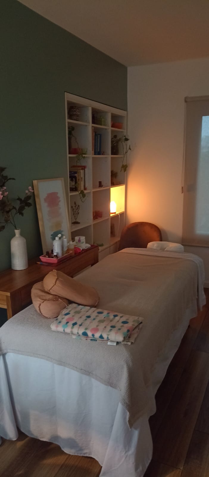 Camilla de masajes con manta beige y almohadones junto a una pared verde salvia